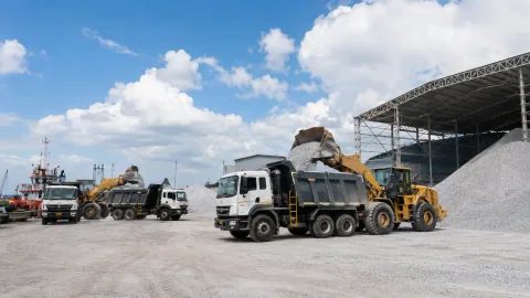 Wheel loaders filling tipper trucks with aggregate beside warehouse sheds